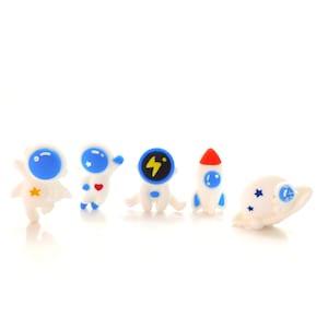 May include: A collection of five space-themed figurines. The set includes two astronauts, a rocket, and a planet with stars. The figurines are white with blue and red accents. One astronaut has a yellow star, and another has a red heart.