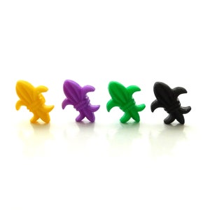 May include: Four fleur-de-lis charms in yellow, purple, green, and black. The charms are made of a shiny material and are arranged in a row against a white background. These charms are often used for jewelry making or craft projects.