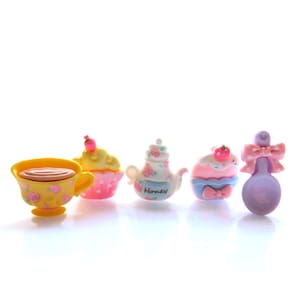 Puede incluir: Colección de miniaturas para una fiesta de té. Incluye una taza amarilla con rosas rosas, dos cupcakes, una tetera con la palabra "Honey" y una cuchara morada con un lazo rosa. Los artículos son de colores brillantes.