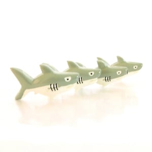 Puede incluir: Cuatro pequeñas figuritas de tiburón de cerámica en fila, todas mirando en la misma dirección. Los tiburones son de color gris claro con vientres blancos y ojos negros. Están dispuestos en línea, con el primer tiburón en la parte posterior y el último tiburón en la parte delantera.