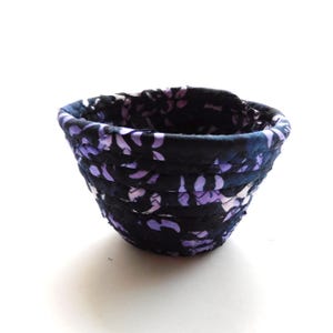 May include: A small, round fabric basket with a floral pattern in shades of purple and white against a dark blue background. The basket is made of tightly coiled fabric, creating a textured appearance. The interior is also dark blue.