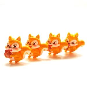 May include: Four orange squirrel figurines with white bellies and rosy cheeks are arranged in a row. Each squirrel is holding a brown acorn. The figurines have large, expressive eyes and are set against a white background.