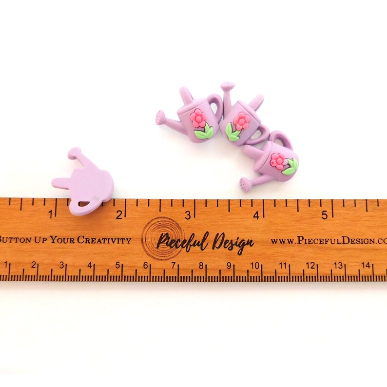 May include: Several small, lavender watering can embellishments with pink flower and green leaf accents. A wooden ruler with inch measurements is in the foreground. The text "BUTTON UP YOUR CREATIVITY" and "Pieceful Design" are visible.