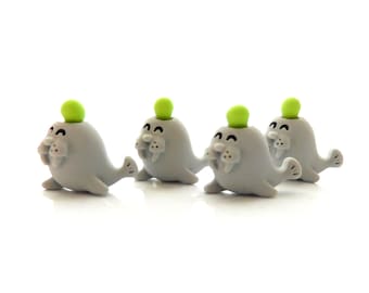 May include: Four small, grey walrus-shaped figurines arranged in a row. Each walrus features a smooth, rounded body, two white tusks, black eyes, and a small, bright lime green ball-shaped accent protruding from the top of its head.