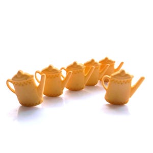 May include: A collection of six small, light orange teapots. Each teapot has a curved spout, a handle, and a decorative pattern around the rim. The teapots are arranged in a row, with a white background.