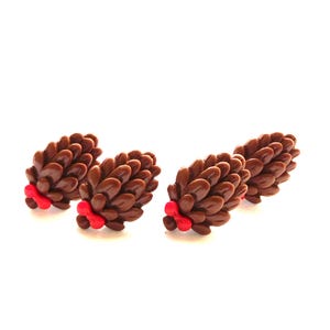 May include: Four brown pine cone shaped ornaments with red accents. The ornaments are arranged in a row on a white background.