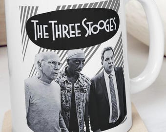 May include: White ceramic mug featuring a black and white image of three men. The text "THE THREE STOOGES" is displayed in a black oval. The mug has a white handle and is placed on a beige coaster.
