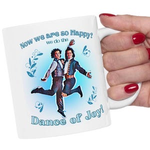 May include: White ceramic mug featuring a colorful image of two men dancing, with the text "Now we are so Happy? we do the Dance of Joy!" in blue. The mug has a white handle and is held by a hand with red nail polish.