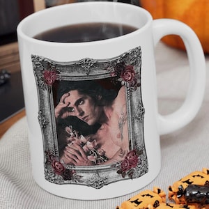 May include: White ceramic mug featuring a framed portrait of a person with long dark hair, holding flowers. The portrait is surrounded by a decorative silver frame with red roses. The mug is filled with a dark liquid and has a white handle.