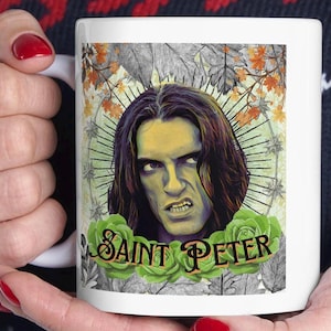 May include: White ceramic mug featuring a portrait of a man with long hair and a stern expression. The mug has the text "Saint Peter" surrounded by green roses. The background includes fall leaves and a halo design.