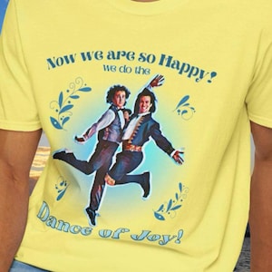 May include: Yellow t-shirt featuring a graphic of two men dancing, with the text "Now we are so Happy! we do the Dance of Joy!" in blue. The design includes blue floral accents. The shirt is made of a soft material.