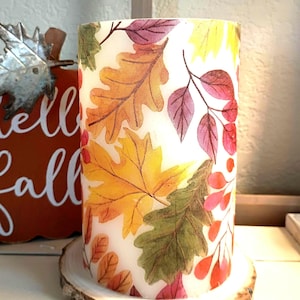 May include: A cylindrical candle featuring a fall leaf design in shades of yellow, orange, red, and green. The candle sits on a round, natural wood slice base, with a rustic bark edge. The background includes a decorative pumpkin and the words "Hello Fall".
