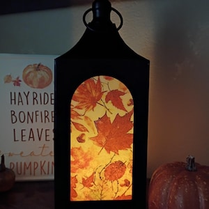 May include: Black metal lantern with a handle, featuring a rectangular window with a fall leaf design. The leaves are in shades of orange and red against a yellow background. A pumpkin and a sign that says "HAY RIDE BONFIRE LEAVES SWEATER PUMPKIN" are in the background.