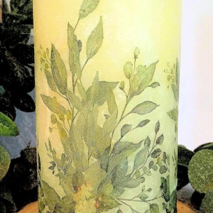 Könnte beinhalten: Eine weiße Säulenkerze mit einem grünen, blättrigen Blumendesign. Die Kerze steht auf einer Holzscheibe.