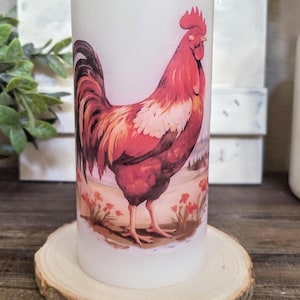 Puede incluir: Una vela cilíndrica blanca con una ilustración detallada de un gallo en tonos rojos, marrones y blancos. La vela está colocada sobre una rodaja de madera redonda y natural. El diseño del gallo incluye un fondo de paisaje.