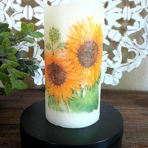 Op de afbeelding: Een witte pilaarvormige kaars met een zonnebloemdessin. De kaars staat op een ronde zwarte voet.