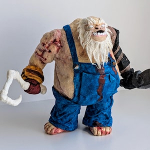 May include: A handmade figurine of a large, white-bearded, monstrous creature wearing blue overalls and holding a hook. The creature has a menacing expression and is covered in dirt and blood.