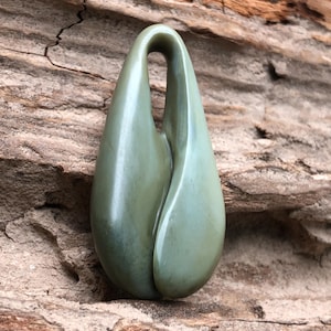 May include: A smooth, green jade pendant with a teardrop shape and a curved top. The polished stone has a glossy finish and is set against a textured, brown wooden background. The pendant is a unique piece of jewelry.