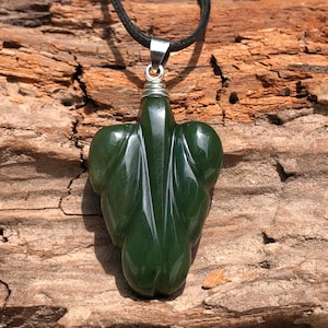Puede incluir: Colgante de jade verde oscuro con forma de hoja. El colgante está suspendido de un collar de cordón negro con una fianza plateada. El jade tiene un acabado pulido y brillante, y el diseño de la hoja presenta vetas detalladas.