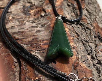 Puede incluir: Un collar con colgante de jade verde oscuro y cordón negro. El colgante tiene forma de punta de flecha lisa y pulida. El collar tiene un cierre plateado y se muestra sobre una superficie marrón texturizada.