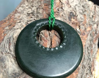 May include: A dark green jade pendant with a round shape and a hole in the center. The pendant is surrounded by a decorative border of small beads.