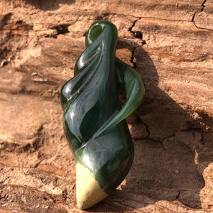 Puede incluir: Un colgante de jade verde oscuro con un diseño orgánico y arremolinado. La piedra pulida tiene un acabado liso y brillante y una base más clara de color crema. La forma del colgante recuerda a una forma fluida.