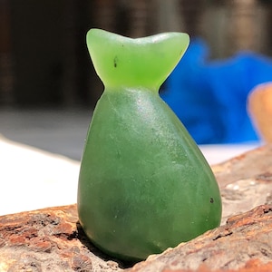 Könnte beinhalten: Eine durchscheinende, grüne, tropfenförmige Skulptur mit ausgestellter Oberseite. Die glatte, polierte Oberfläche des Steins reflektiert das Licht und hebt seine natürliche Farbe hervor. Die Skulptur steht vor einem natürlichen Holzuntergrund.