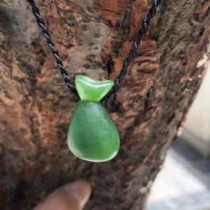 May include: A green jade pendant in the shape of a small pouch, suspended from a black cord. The jade has a smooth, polished surface and a vibrant green color. The cord is a twisted black material.