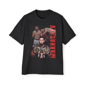 May include: Black t-shirt featuring a UFC fighter holding championship belts. The shirt has a graphic of the fighter and the word "MAKHACHEV" in red vertical text. The design includes action shots of the fighter in the ring.
