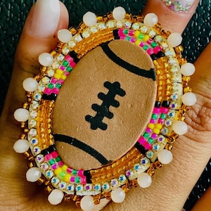 May include: A beaded ring with a brown football design. The ring is made with white, brown, orange, pink, and green beads.