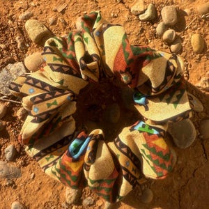 May include: A large, patterned scrunchie with a Southwestern design. The fabric features geometric shapes in shades of brown, red, green, and blue on a tan background. The scrunchie is lying on a bed of red dirt and small rocks.
