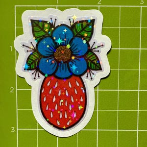 May include: A die-cut sticker featuring a stylized design of a blue flower atop a red strawberry. The flower has a brown center and green leaves. The sticker has a holographic, glittery effect.
