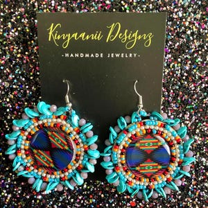 May include: A pair of turquoise and silver earrings with a beaded design and a colorful geometric pattern in the center. The earrings are handmade and feature the text "Kinyaanii Designs - HANDMADE JEWELRY - ".