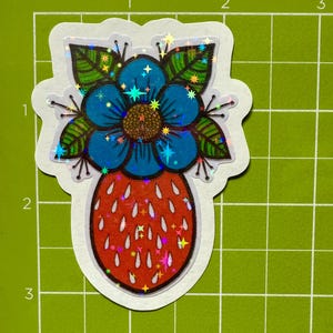 May include: A holographic sticker featuring a blue flower with green leaves and a red strawberry. The sticker has a glittery finish.