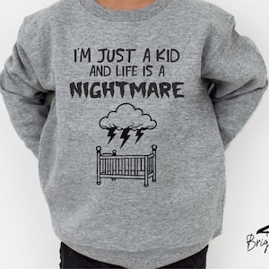 以下が含まれることがあります： 「I'M JUST A KID and LIFE IS A NIGHTMARE」の黒い文字がプリントされたグレーのスウェットシャツ。その下に、雷雲とベビーベッドのグラフィックがあります。Briggs Design Apparelのロゴが下にあります。