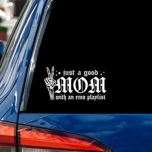 Op de afbeelding: Witte sticker op een blauwe autoruit. De sticker leest "just a good MOM with an emo playlist" in een gotisch lettertype, met een skelet hand die een vredesteken maakt. De auto staat buiten geparkeerd.