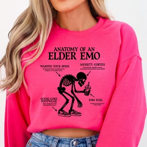 May include: A bright pink sweatshirt with the text "ANATOMY OF AN ELDER EMO" and a black graphic of a skeleton. The graphic includes labeled body parts like "WARPED TOUR SPINE" and "ANXIETY CORTEX". The sweatshirt also features the "Briggs Design Apparel" logo.