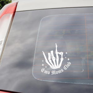 Op de afbeelding: Witte sticker op een autoruit met een skelet hand die de middelvinger opsteekt. De tekst "Emo Moms Club" staat in een cirkel rond de hand. De auto is rood en wit.