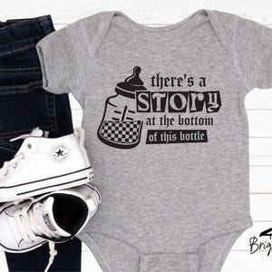 May include: A gray baby onesie with the text "there's a STORY at the bottom of this bottle" and a bottle graphic. The onesie is paired with dark blue jeans and white high-top sneakers.