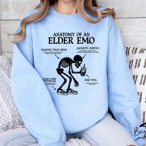 May include: Light blue sweatshirt with the text "ANATOMY OF AN ELDER EMO" and a skeleton graphic. The graphic includes labeled body parts like "WARPED TOUR SPINE" and "ANXIETY CORTEX". The sweatshirt is a casual style.