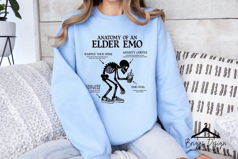 May include: Light blue sweatshirt with the text "ANATOMY OF AN ELDER EMO" and a skeleton graphic. The graphic includes labeled body parts like "WARPED TOUR SPINE" and "ANXIETY CORTEX". The sweatshirt is a casual style.