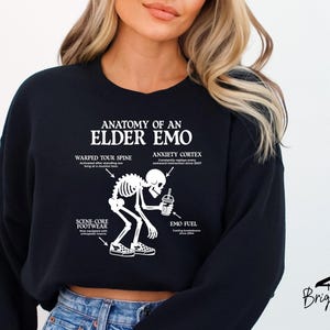 May include: Black sweatshirt with the text "ANATOMY OF AN ELDER EMO" and a skeleton graphic. The graphic includes labels for "WARPED TOUR SPINE", "ANXIETY CORTEX", "SCENE CORE FOOTWEAR", and "EMO FUEL". The sweatshirt is from Briggs Design Apparel.