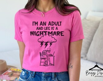 May include: A pink t-shirt with the phrase "I'M AN ADULT AND LIFE IS A NIGHTMARE" in black text. Below the text is a graphic of a cloud with lightning bolts and a skeleton sitting at a desk. The shirt is a crew neck.