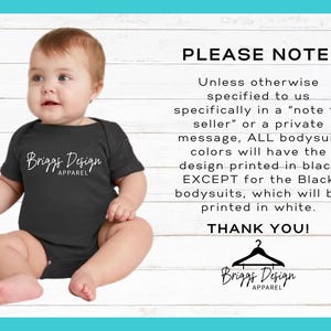 May include: A black baby bodysuit with the text "Briggs Design Apparel" in white script. The image includes a baby sitting on a white wooden background with text that says "PLEASE NOTE:" and other details about the product.