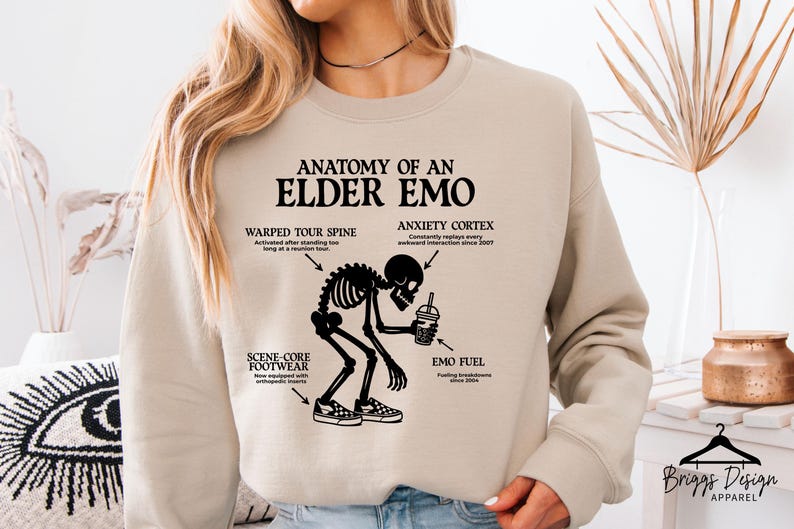 May include: Beige sweatshirt with the text "ANATOMY OF AN ELDER EMO" and a black skeleton graphic. The graphic includes labels for "WARPED TOUR SPINE", "ANXIETY CORTEX", "SCENE-CORE FOOTWEAR", and "EMO FUEL".