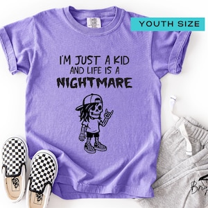 May include: A lilac youth-sized t-shirt with the text "I'M JUST A KID AND LIFE IS A NIGHTMARE" and a skeleton graphic. The image includes checkered slip-on shoes and gray sweatpants.