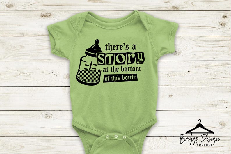 May include: Light green baby onesie with a black graphic of a baby bottle and the text "there's a STORY at the bottom of this bottle." The onesie has short sleeves and snaps at the bottom. Briggs Design Apparel logo is at the bottom.