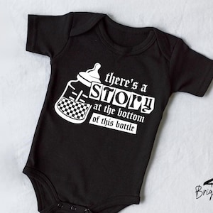 May include: Black baby onesie with white text and a bottle graphic. The text reads "there's a STORY at the bottom of this bottle." The bottle graphic has a checkerboard pattern. The onesie is short-sleeved.
