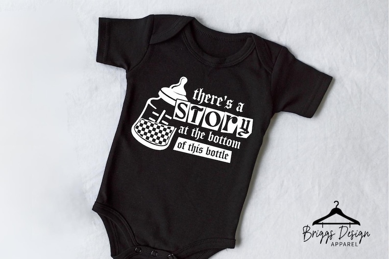 May include: Black baby onesie with white text and a bottle graphic. The text reads "there's a STORY at the bottom of this bottle." The bottle graphic has a checkerboard pattern. The onesie is short-sleeved.
