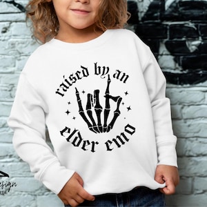 May include: White sweatshirt with the text "raised by an elder emo" in a gothic font, featuring a black skeleton hand gesture. The design includes star accents. The brand logo is at the bottom.